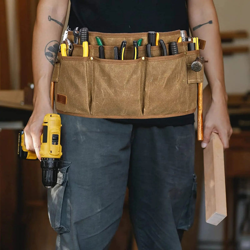 Person wearing a tool belt with various tools and holding a drill and piece of wood.