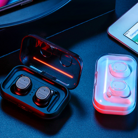 Wireless earbuds in open and closed cases on a dark surface with a phone and tablet in the background.
