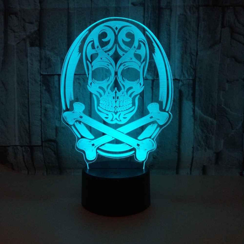 A 3D optical illusion LED night light featuring a glowing skull and crossbones design, illuminated in a bright cyan-blue light on a dark base, set against a dark textured stone wall background.