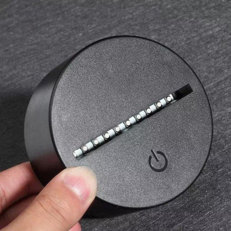 A close-up of a round black electronic device with a textured surface, featuring a diagonal strip of white LED light elements and a power symbol icon. A person's thumb is visible holding the device on a dark gray textured background.