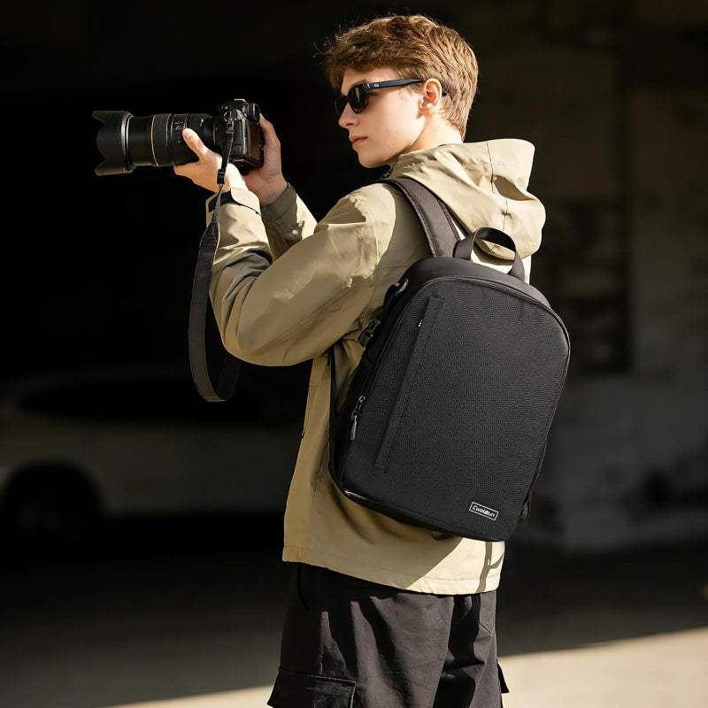 Person holding a camera with a black backpack, standing outdoors.
