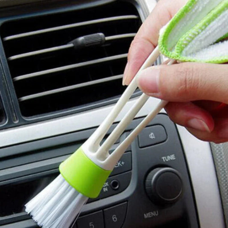 Car air vent cleaning brush with green handle held by a hand in front of a car's air vent.