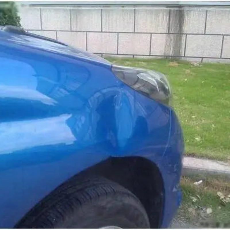 Blue car with a dent on its side next to a grassy area and wall.
