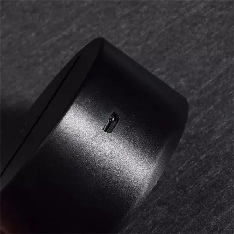 Close-up of a matte black cylindrical product, showing a small recessed slot or button on its side, photographed against a dark textured background.