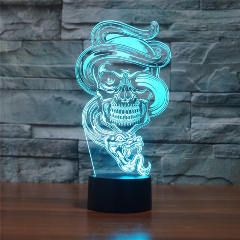 An LED acrylic 3D optical illusion night light featuring a glowing skull wearing a hat and a snake in the foreground, set on a dark base against a gray stone wall background.
