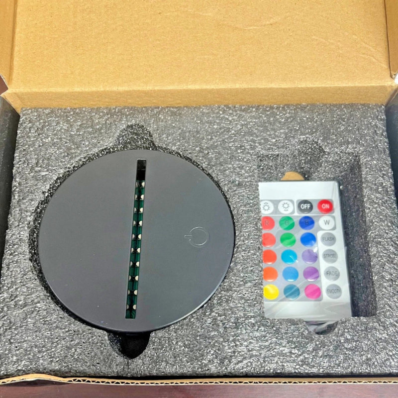 A round black LED light strip fixture with a vertical slot, shown inside its open cardboard box next to a white remote control with colorful buttons, set in gray foam packaging.